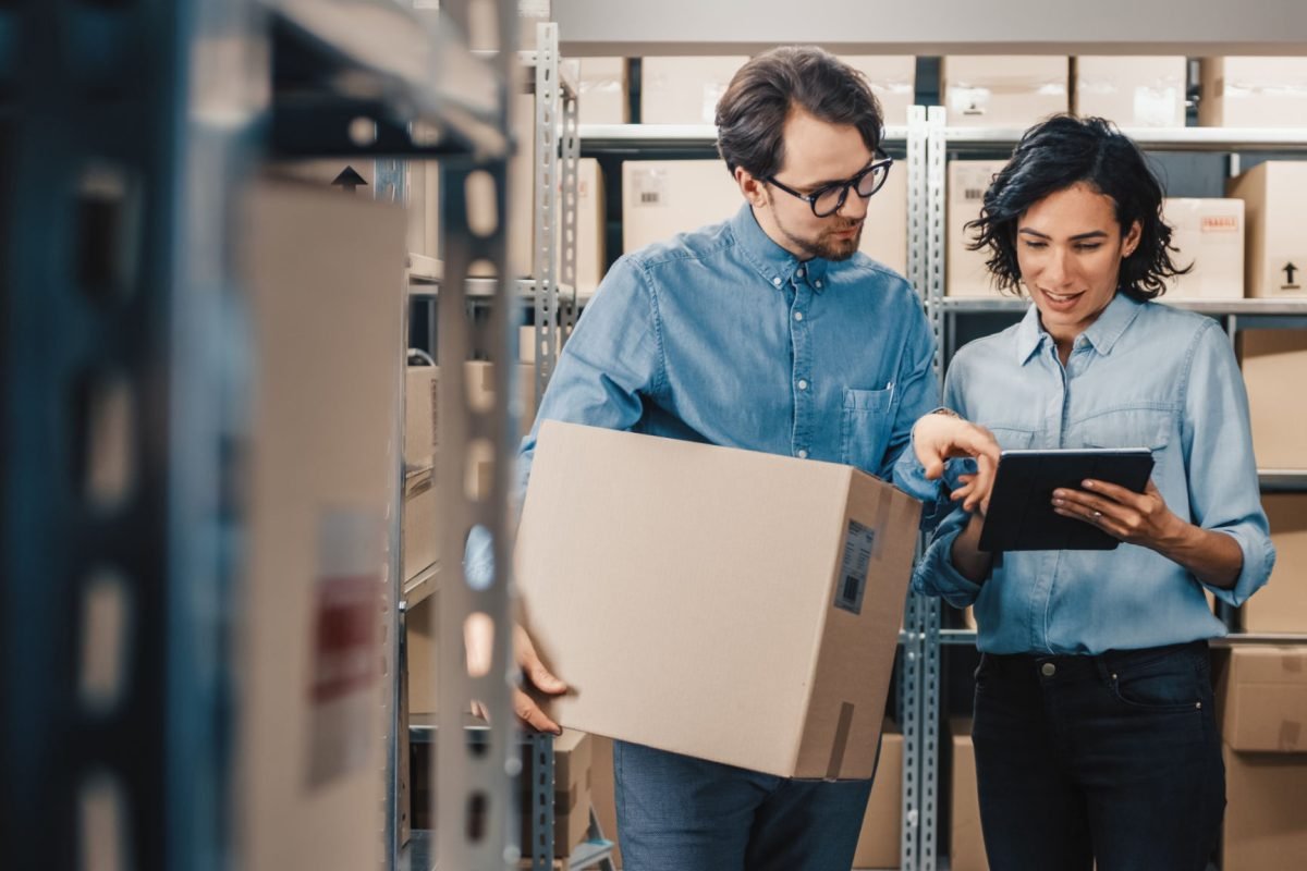 Female Inventory Manager Shows Digital Tablet Information to a Worker Holding Cardboard Box, They Talk and Do Work. In the Background Stock of Parcels with Products Ready for Shipment.