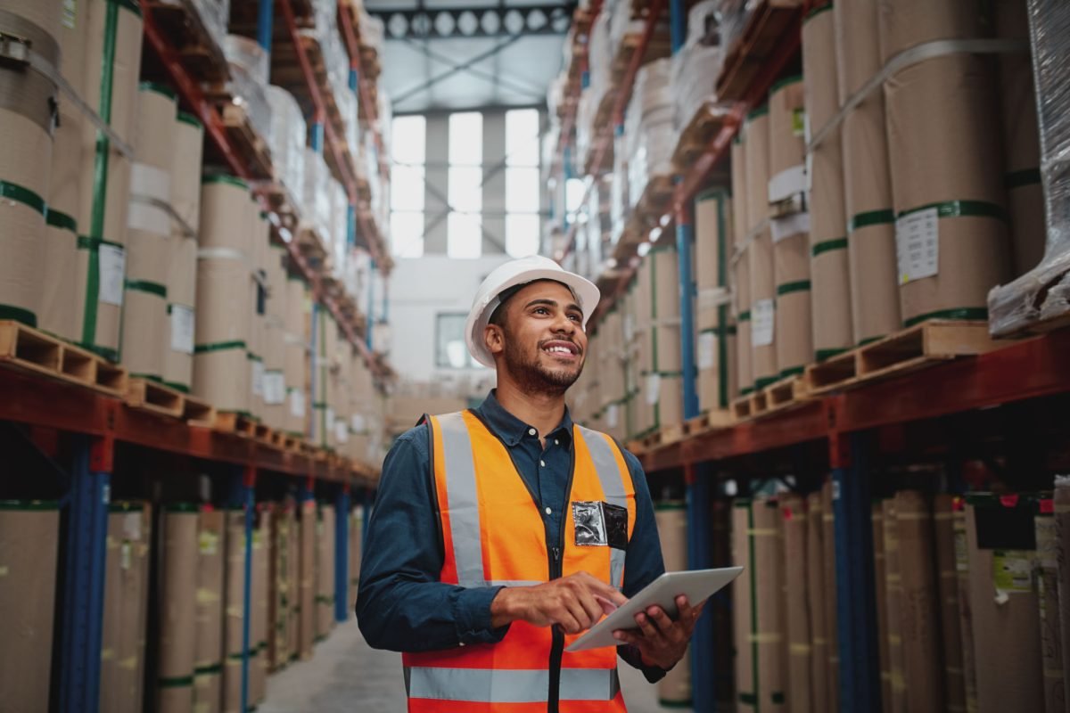 Portrait of young man wearing safety jacket holding digital tablet standing in factory warehouse