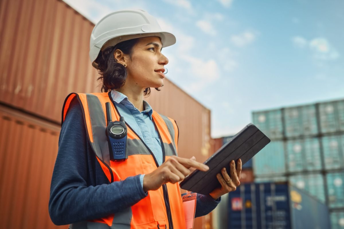 Smiling Portrait of a Beautiful Latin Female Industrial Engineer in White Hard Hat, High-Visibility Vest Working on Tablet Computer. Inspector or Safety Supervisor in Container Terminal.