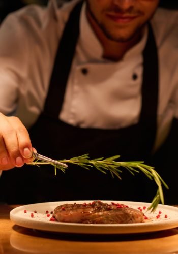 Hand of young male chef putting aromatic herb on top of roasted meat seasoned with spices while preparing tasty dish for serving