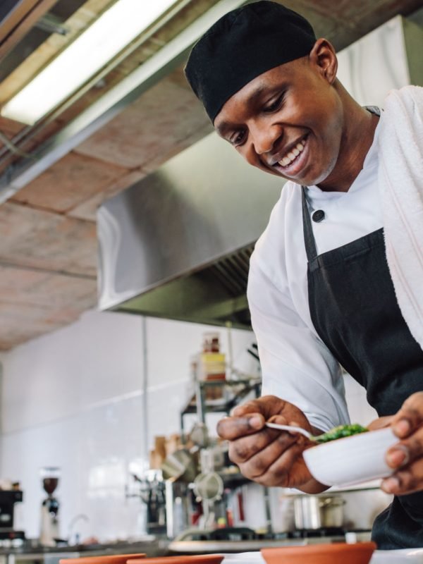 Gourmet chef in uniform cooking in a commercial kitchen. Happy male cook wearing apron standing by kitchen counter preparing food.